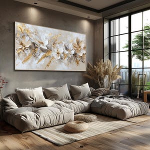 Op de afbeelding: Een groot abstract schilderij met witte bloemen en gouden accenten hangt boven een beige hoekbank in een moderne woonkamer. De bank heeft een gestoffeerd ontwerp en is L-vormig geplaatst. De kamer heeft een groot raam met uitzicht op een groene boom en een skyline.