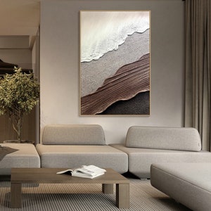May include: Abstract painting with textured layers of white, brown, and black paint. The painting is framed in a thin gold frame and hangs on a wall in a modern living room.