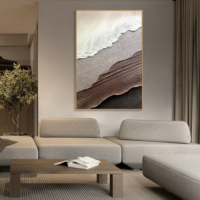 May include: Abstract painting with textured layers of white, brown, and black paint. The painting is framed in a thin gold frame and hangs on a wall in a modern living room.