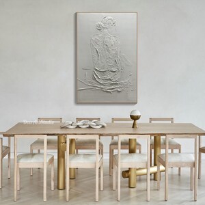 Puede incluir: Una pintura blanca texturizada de una persona sentada de espaldas al espectador. La pintura est&aacute; colgada en una pared blanca sobre una mesa de comedor de madera con seis sillas.