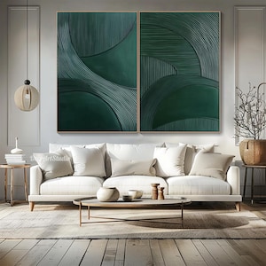 May include: Two-panel abstract wall art featuring a textured, dark green design. The artwork has a series of curved lines and shapes, creating a sense of depth and movement. The art is framed in a light-colored wood and hangs above a white sofa.