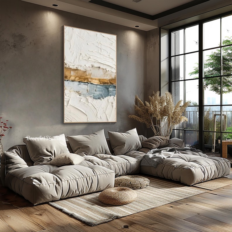 May include: An abstract textured painting in a gold frame, featuring white, gold, and blue horizontal bands. The artwork hangs above a large, modular beige sofa with several pillows. The room has a modern design with a large window.