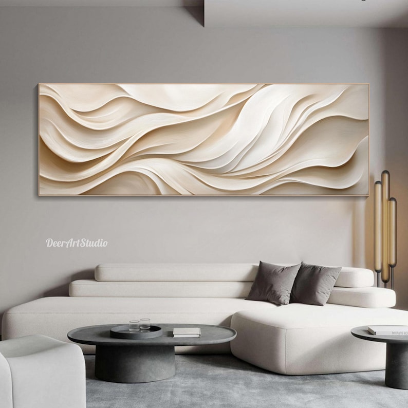 May include: Abstract wall art featuring a beige and white wavy pattern. The artwork is framed in a thin gold frame and is hanging on a white wall above a white couch.