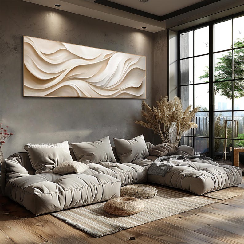 May include: A large, abstract painting with white and beige waves hangs above a sectional sofa with plush cushions. The sofa is in a modern living room with large windows and a view of trees.