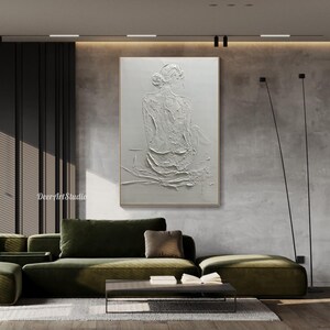 Puede incluir: Pintura abstracta texturizada blanca de una persona sentada de espaldas al espectador. La pintura est&aacute; enmarcada y colgada en una pared de una sala de estar moderna.