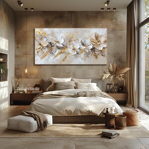 Op de afbeelding: Een slaapkamerinterieur met een groot abstract schilderij van witte bloemen met gouden accenten boven een bed met witte beddengoed en bruine kussens. De kamer heeft een bruin tapijt, een witte poef en twee kleine houten bijzettafels met decoratieve items.