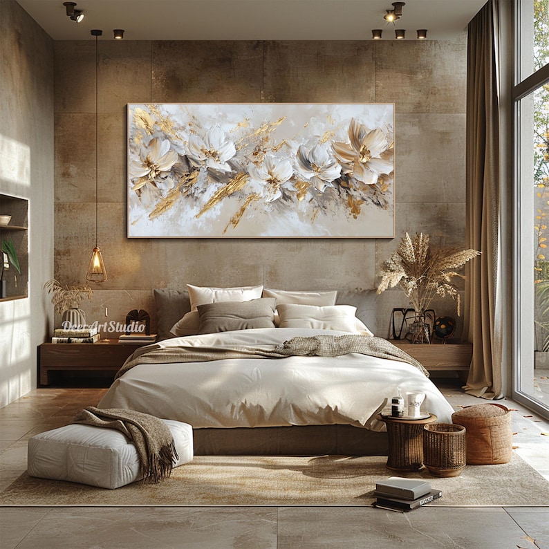 Op de afbeelding: Een slaapkamerinterieur met een groot abstract schilderij van witte bloemen met gouden accenten boven een bed met witte beddengoed en bruine kussens. De kamer heeft een bruin tapijt, een witte poef en twee kleine houten bijzettafels met decoratieve items.