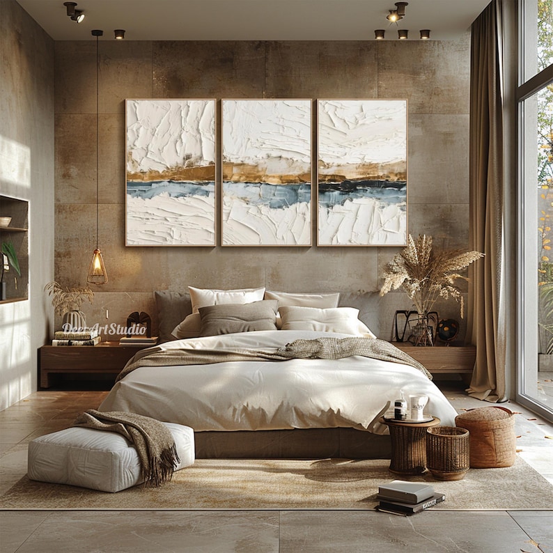 May include: A bedroom with a neutral color palette, featuring a bed with beige bedding and decorative pillows. Above the bed, three framed abstract paintings in white, gold, and blue tones hang on the wall. The room includes a rug, side tables, and a large window.