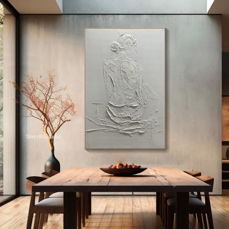 Puede incluir: Pintura abstracta texturizada blanca de una persona sentada de espaldas al espectador. La pintura est&aacute; sobre un lienzo blanco y est&aacute; enmarcada en un fino marco dorado. La pintura est&aacute; colgada en una pared blanca en una habitaci&oacute;n con una mesa y sillas de madera.