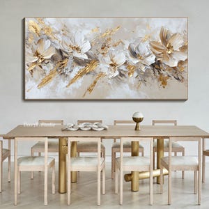 Op de afbeelding: Abstract schilderij met witte en gouden bloemen op een beige achtergrond. Het schilderij hangt boven een houten eettafel met stoelen.