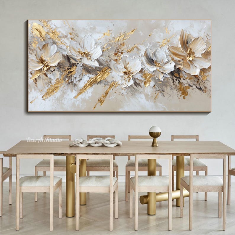 Op de afbeelding: Abstract schilderij met witte en gouden bloemen op een beige achtergrond. Het schilderij hangt boven een houten eettafel met stoelen.