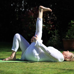 May include: A person in white clothing is lying on a green yoga mat on a grassy lawn, performing a yoga stretch. The person is holding their leg up in the air, with their foot flexed. The background includes trees and a potted plant.
