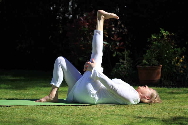 May include: A person in white clothing is lying on a green yoga mat on a grassy lawn, performing a yoga stretch. The person is holding their leg up in the air, with their foot flexed. The background includes trees and a potted plant.