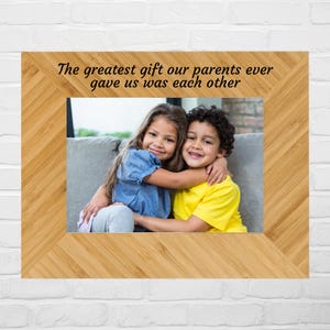 Puede incluir: Un marco de fotos de madera con un acabado marrón claro y un borde biselado. El marco tiene una foto de dos niños abrazándose, con el texto "El mejor regalo que nuestros padres nos dieron fue el uno al otro" inscrito en el marco.