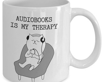 May include: White ceramic mug with a black and white illustration of a cat wearing headphones and listening to an audiobook. The text "Audiobooks is my therapy" is printed on the mug.