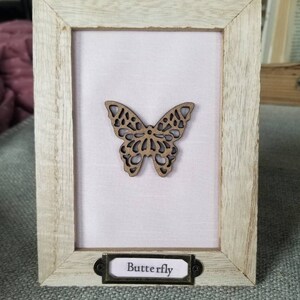 Puede incluir: Un marco de madera con un fondo rosa claro y un recorte de mariposa marrón. El marco tiene una etiqueta de metal con la palabra "Butterfly" en ella.