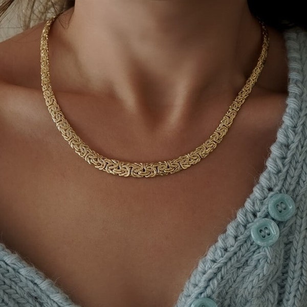 Pode incluir: Um colar de corrente de ouro com um design de estilo bizantino. O colar é usado por uma pessoa com um suéter azul claro.