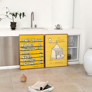 May include: Two framed illustrations on a white wall. The illustration on the left is a yellow and black coffee bar menu with prices listed. The illustration on the right is a yellow and black drawing of a coffee grinder with the text "It's a grind set" and "You wouldn't understand..."