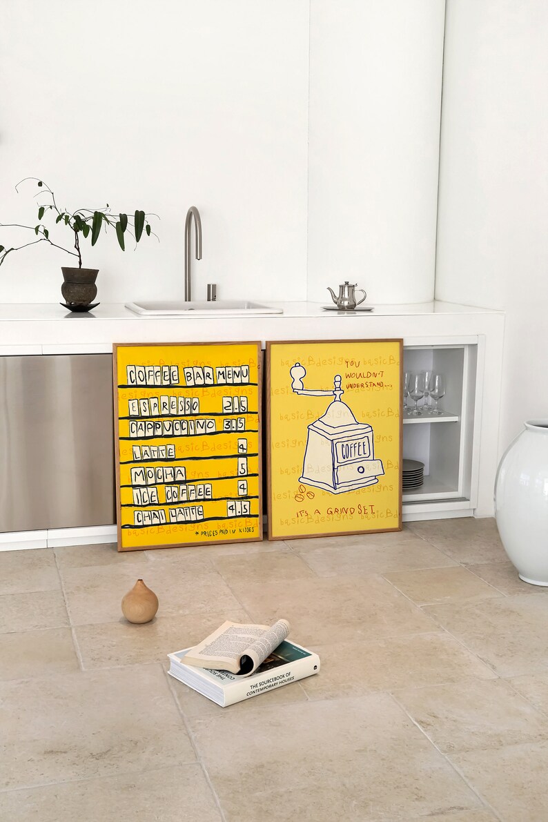 May include: Two framed illustrations on a white wall. The illustration on the left is a yellow and black coffee bar menu with prices listed. The illustration on the right is a yellow and black drawing of a coffee grinder with the text "It's a grind set" and "You wouldn't understand..."