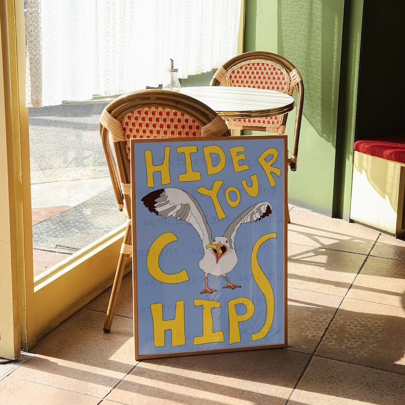 May include: A blue and yellow sign with the text "Hide Your Chips" and a cartoon seagull with a beak open wide. The sign is leaning against a wooden chair in a cafe setting.