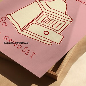 May include: A pink poster with a red illustration of a coffee grinder with the text "COFFEE" on it. The text "GRIND SET" is written in red at the bottom of the poster.