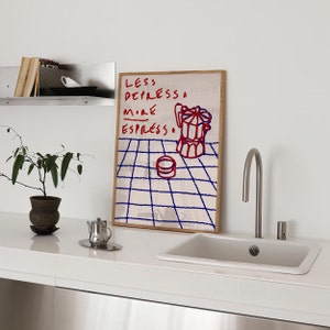 May include: A framed print with a white background and blue grid pattern. The print features red and blue text that reads "Less Depresso More Espresso." A red and white illustration of a coffee pot is on the print.