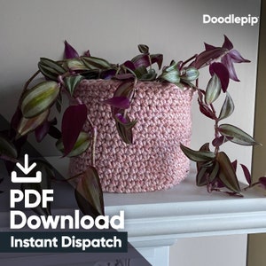 May include: A pink crocheted plant pot with a leafy green and purple plant inside. The pot is sitting on a white shelf. The text "PDF Download Instant Dispatch" is below the pot.