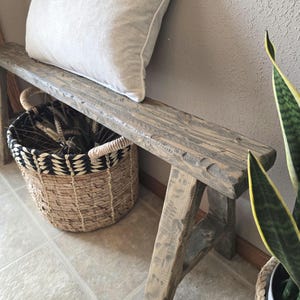 May include: A rustic wooden bench with a distressed finish. A light-colored pillow rests on the bench. A woven basket with a black and white pattern and a plant in a white pot are nearby. The bench is made of wood.