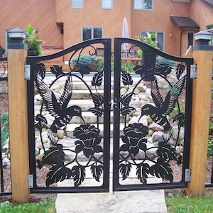 May include: A black metal gate with a decorative design featuring hummingbirds and flowers. The gate is set against a backdrop of a brown wooden fence and a green lawn.