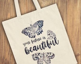 Puede incluir: Bolso tote de color crema con tres ilustraciones detalladas de mariposas en tonos azules y grises. La frase "your future is beautiful" está escrita en una elegante tipografía. El bolso tiene dos asas color crema.
