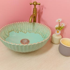 May include: A mint green ceramic vessel sink with a scalloped edge and a gold faucet. The sink has a light blue interior and a decorative drain. A small vase with white flowers and a gray candle holder are on the counter.