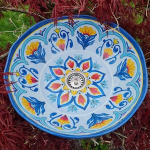 Könnte beinhalten: Ein runder, handbemalter Keramikwaschtisch mit einem lebendigen Blumendesign. Das Waschbecken hat einen hellblauen Hintergrund mit gelben, roten und blauen Blumenmustern. Der Rand ist tiefblau und der Abfluss ist silberfarben.