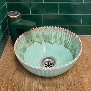 May include: A round, fluted ceramic vessel sink with a light mint green interior and a darker green, textured rim featuring reddish-brown accents. The sink includes a silver metal drain and is paired with a dark green faucet with copper trim.