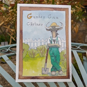 Puede incluir: Una ilustración de acuarela de un ñu de dibujos animados con un sombrero de paja y un mono, sosteniendo una pala. El ñu está de pie en un jardín con una valla blanca de madera y arbustos verdes. El texto "Gunter Gnu Gärtner" está escrito encima del ñu.