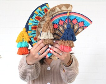 May include: A collection of colorful Wayúu clutch bags with fringe tassels. The bags are made of woven fabric and feature geometric patterns in shades of blue, red, yellow, and orange.