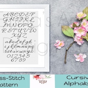 May include: A framed cross-stitch pattern featuring a cursive alphabet and numbers in black thread on white fabric. The framed artwork is displayed on a gray surface, accompanied by pink flowers and green leaves. The text at the bottom reads "Cross-Stitch Pattern" and "Cursive Alphabet."