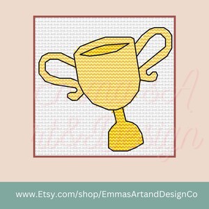 May include: A cross-stitch design of a gold trophy on a white background, framed in a red border. The trophy has a cup-shaped top with two handles and a pedestal base. The website address www.Etsy.com/shop/EmmasArtandDesignCo is at the bottom.