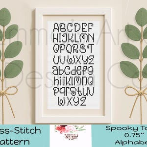 May include: A framed cross-stitch pattern featuring the alphabet in a black, blocky font. The framed artwork is white and is flanked by green leaf sprigs tied with twine. The text "Spooky Town 0.75" Alphabet" is visible.