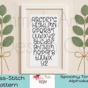 May include: A framed cross-stitch alphabet pattern in black thread. The letters are displayed in uppercase and lowercase. The frame is white, and the background is a light beige. Green leaf stems with twine bows flank the frame.