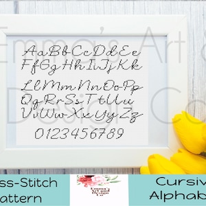 May include: A framed cross-stitch pattern featuring a cursive alphabet and numbers in black thread. The white frame sits against a light wood background, with yellow tulips in the corner. The text "Cross-Stitch Pattern" and "Cursive Alphabet" are at the bottom.
