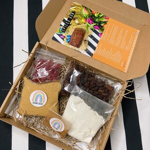 May include: A brown cardboard box filled with ingredients for a spiced bun recipe. The box contains a bag of brown sugar, a bag of flour, a bag of raisins, a bag of candied cherries, and a small bag of spices. The box also includes a recipe card with the title "Spiced Bun".