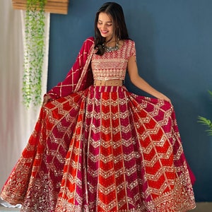 以下が含まれることがあります： 赤い刺繍の入ったピンクのレヘンガを着た女性。レヘンガは、シェブロン柄の重ね着のスカートと、それに合わせた刺繍の入ったブラウスを備えています。