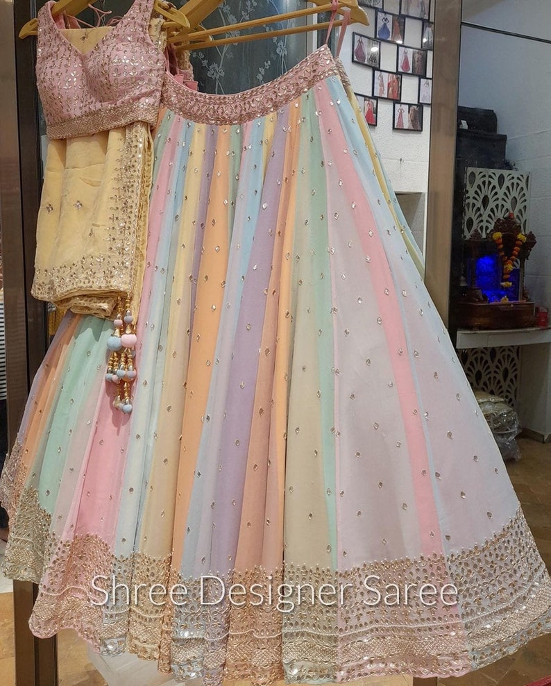 May include: A rainbow-coloured skirt with a gold embroidered border and a matching pink blouse. The skirt is made of a sheer fabric and has a flowing silhouette. The text "Shree Designer Saree" is visible at the bottom of the skirt.