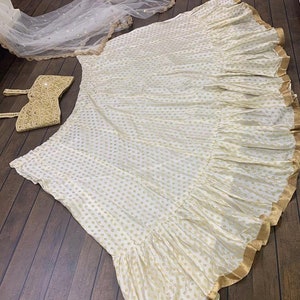 May include: A white lehenga with gold embroidery and a gold trim. The lehenga has a ruffled skirt and is paired with a matching white crop top.