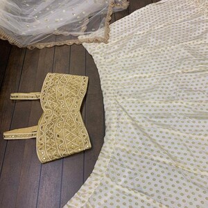 May include: A white skirt with gold polka dots, a matching white and gold embroidered crop top, and a white sheer scarf with gold sequins.