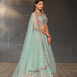 May include: A mint green lehenga with floral embroidery and a matching dupatta. The lehenga features intricate patterns and red and pink floral accents along the hem. The text "Blacknooz" is visible on the skirt.