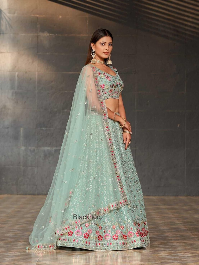 May include: A mint green lehenga with floral embroidery and a matching dupatta. The lehenga features intricate patterns and red and pink floral accents along the hem. The text "Blacknooz" is visible on the skirt.
