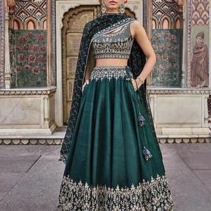 Puede incluir: Un lehenga verde oscuro, una prenda tradicional india, con intrincados bordados dorados en el dobladillo y el corpi&ntilde;o. La parte superior sin mangas est&aacute; adornada con detalles plateados. Una chal a juego cae sobre los hombros. El atuendo se usa frente a un fondo arquitect&oacute;nico ornamentado.