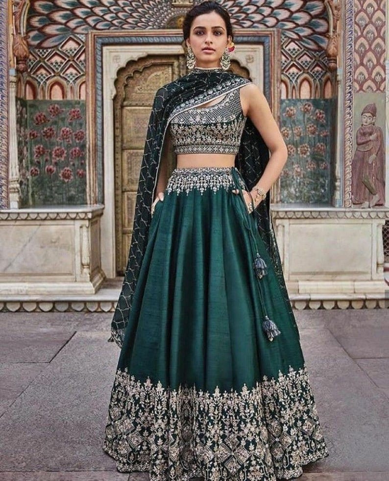 Puede incluir: Un lehenga verde oscuro, una prenda tradicional india, con intrincados bordados dorados en el dobladillo y el corpi&ntilde;o. La parte superior sin mangas est&aacute; adornada con detalles plateados. Una chal a juego cae sobre los hombros. El atuendo se usa frente a un fondo arquitect&oacute;nico ornamentado.