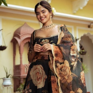 May include: A woman wearing a black and brown floral print lehenga with a gold trim. She is wearing a gold necklace and earrings. The background is a yellow wall.